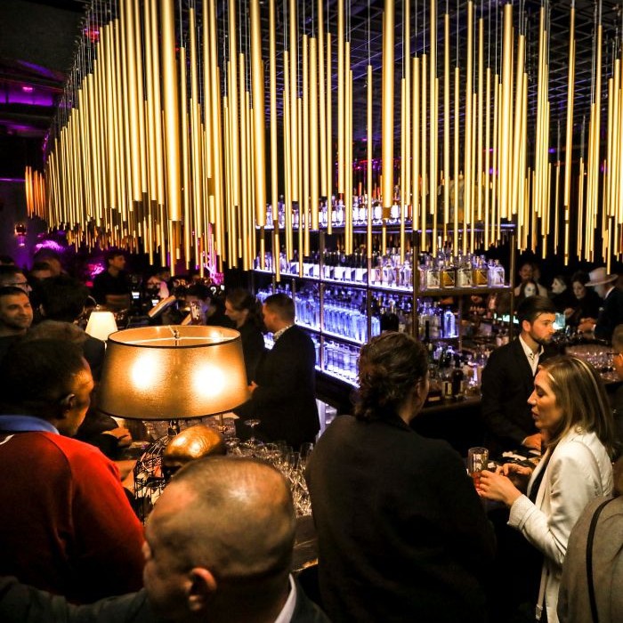 Lebendige Atmosphäre in der Gin-Bar House of Gin im Hotel Palace Berlin, mit goldenen Stangen an der Decke, einem Barkeeper hinter der Bar, bunten Lichtern und Live-Musik. Gäste genießen gesellige Gespräche.