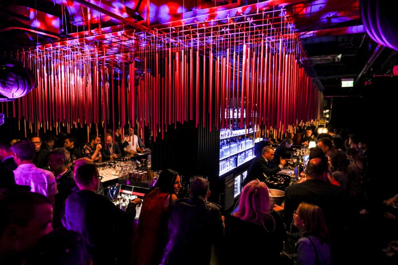 Vibrant atmosphere at House of Gin bar in Hotel Palace Berlin, with red rods hanging from the ceiling, a bartender serving drinks, colorful lighting, and live music. Guests enjoy lively conversations.