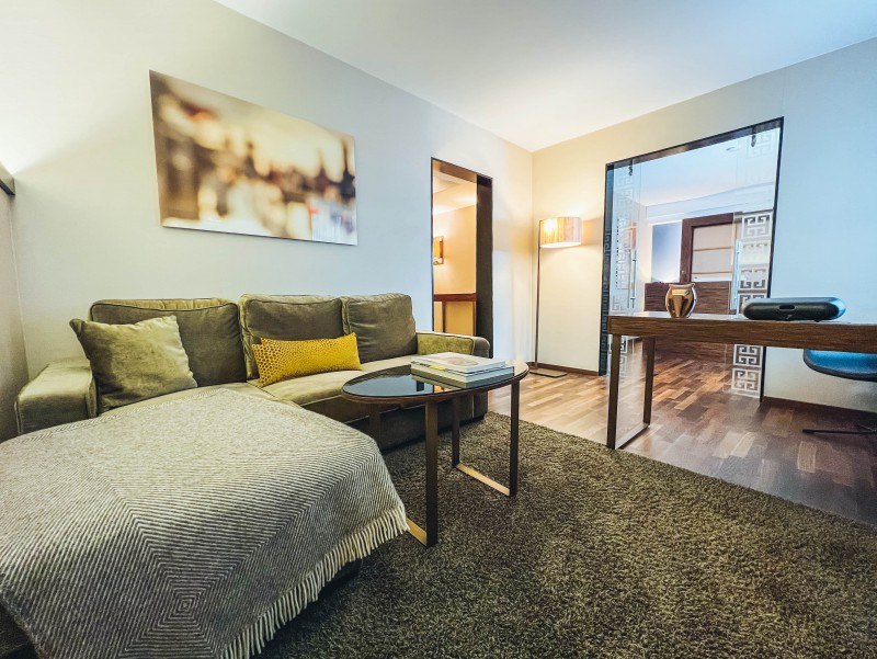 Die Suite im Hotel Palace Berlin kombiniert Wohn-, Arbeits- und Schlafbereich in einem stilvollen Ambiente. Mit Sofa, Schreibtisch, Fernseher und dekorativen Pflanzen bietet sie Komfort, während warmes Licht und Holzboden für Gemütlichkeit sorgen.