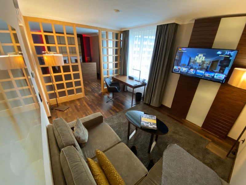 Die Suite im Hotel Palace Berlin kombiniert Wohn-, Arbeits- und Schlafbereich in einem stilvollen Ambiente. Mit Sofa, Schreibtisch, Fernseher und dekorativen Pflanzen bietet sie Komfort, während warmes Licht und Holzboden für Gemütlichkeit sorgen.