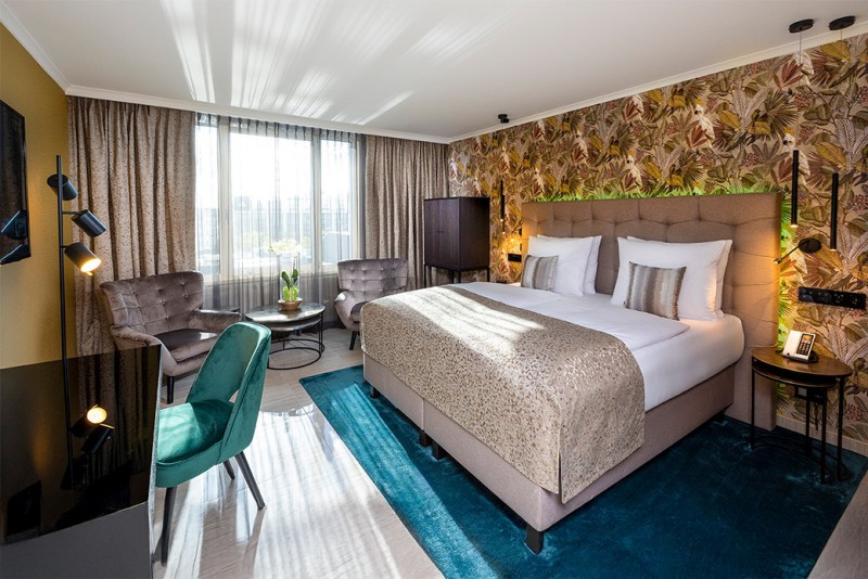 Business-Premium-Zimmer im Hotel Palace Berlin mit großem Bett, blumiger Tapete, Sessel, grauer Couch und Tageslicht durch transparente Vorhänge.