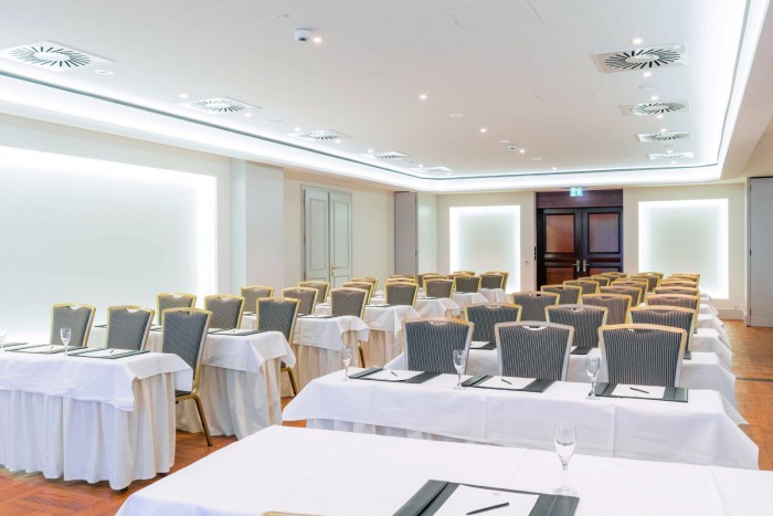 Heller, leerer Tagungsraum im Hotel Palace Berlin mit Reihen von Stühlen vor einer Projektionsleinwand. Weiß gedeckte Tische sind mit Notizblöcken, Stiften und Weingläsern ausgestattet. Holzboden und modernes Deckendesign ergänzen die Ausstattung.