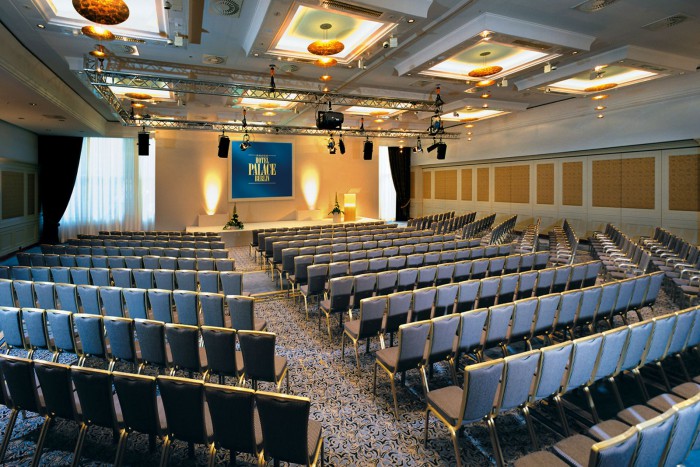 Moderner Konferenzraum im Hotel Palace Berlin mit flexiblen Sitzgelegenheiten, eleganten grauen Stühlen, einer Bühne mit Podium und Leinwand in einem gut beleuchteten, mit Teppich ausgelegten Raum.