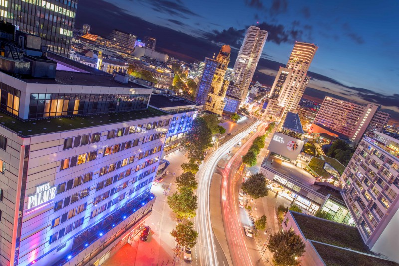 Nighttime cityscape in Berlin with illuminated buildings, busy street, car light trails, and the Kaiser Wilhelm Memorial Church near Hotel Palace Berlin.