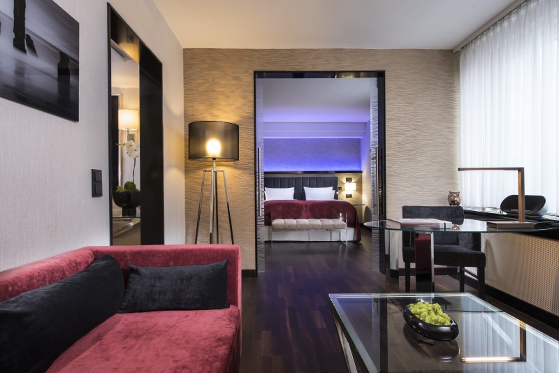 Die Grand Suite im Hotel Palace Berlin vereint modernes Design mit Komfort. Eine rote Couch, ein Glascouchtisch und eine Stehlampe setzen stilvolle Akzente, während das Schlafzimmer in neutralen Tönen und sanfter Beleuchtung Ruhe ausstrahlt. Der elegant
