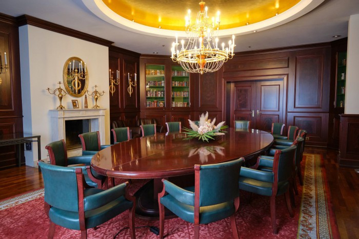 Das luxuriöse Kaminzimmer im Tagungsbereich des Hotel Palace Berlin mit ovalem Holztisch, umgeben von grünen Stühlen. Ein Kronleuchter ziert die Decke, während Holzvertäfelung, Bücherregale und ein Kamin eine elegante Atmosphäre schaffen.