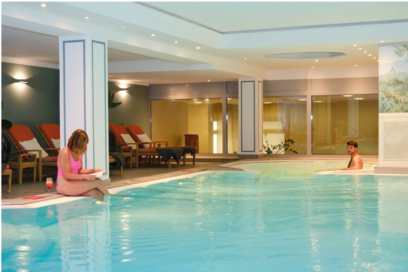 Two people relax at the indoor pool of Palace Spa & Fitness at Hotel Palace Berlin. One is reading by the poolside, while the other enjoys the water. Lounge chairs line the background of this tranquil oasis.