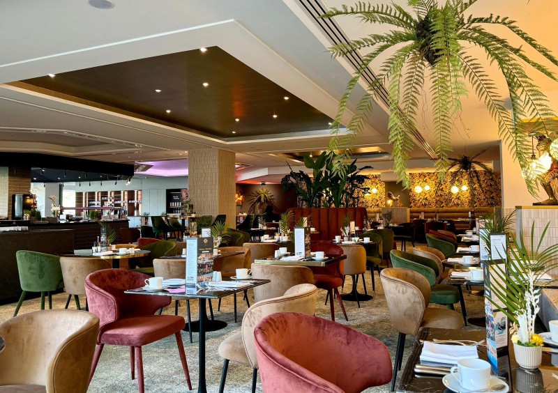 Modern restaurant interior at The Food Court breakfast restaurant in Hotel Palace Berlin. Set tables, colorful chairs, patterned wallpaper, and abundant greenery with hanging ferns and potted plants create an inviting atmosphere.