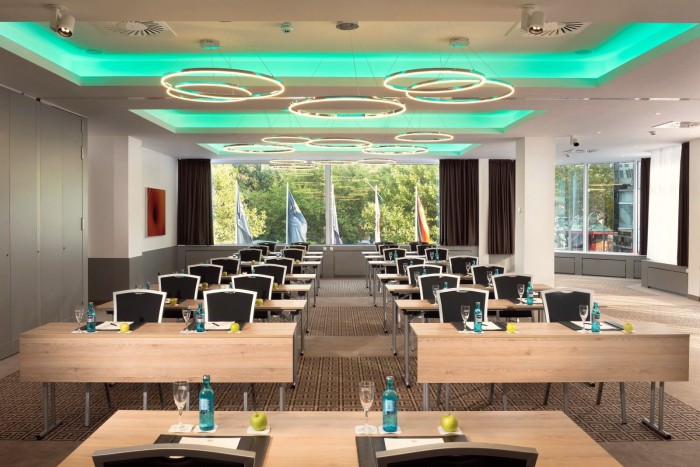Moderner Tagungsraum im Hotel Palace Berlin mit Holztischen und schwarzen Stühlen in Reihen. Jeder Tisch ist mit Wasserflaschen, Notizblöcken und grünen Äpfeln ausgestattet. Runde Deckenleuchten und türkisfarbene Beleuchtung ergänzen die Ausstattung